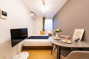 A compact hotel room with a bed, a flat-screen TV, a small dining table with plates and a cup, and a framed picture on the wall. - Maison Ash Ikebukuro by Tranova (Tokyo)
