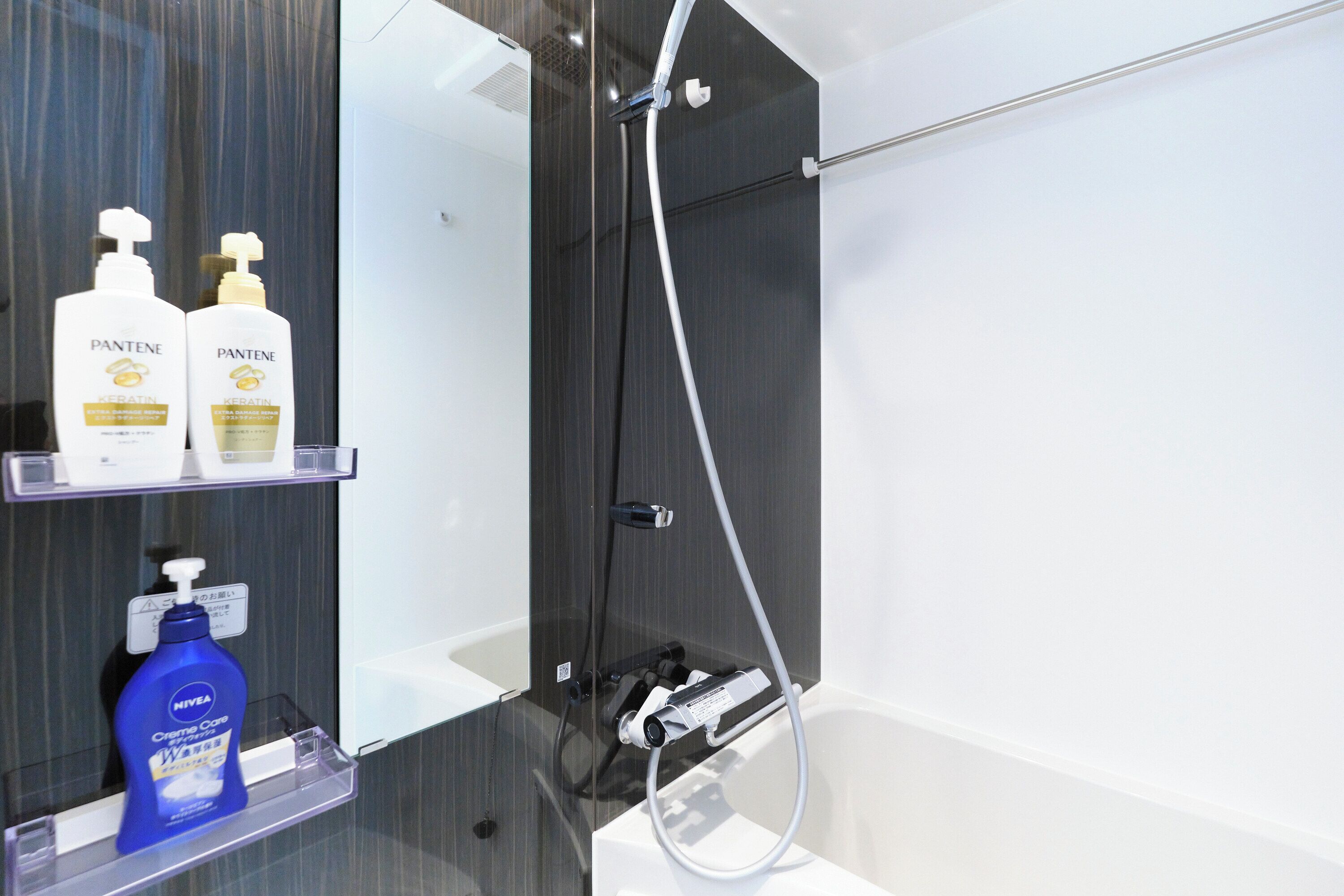 A modern bathroom with a glass shower enclosure, a white bathtub, and a shelf with two Pantene shampoo bottles and a Nivea Care lotion bottle.
