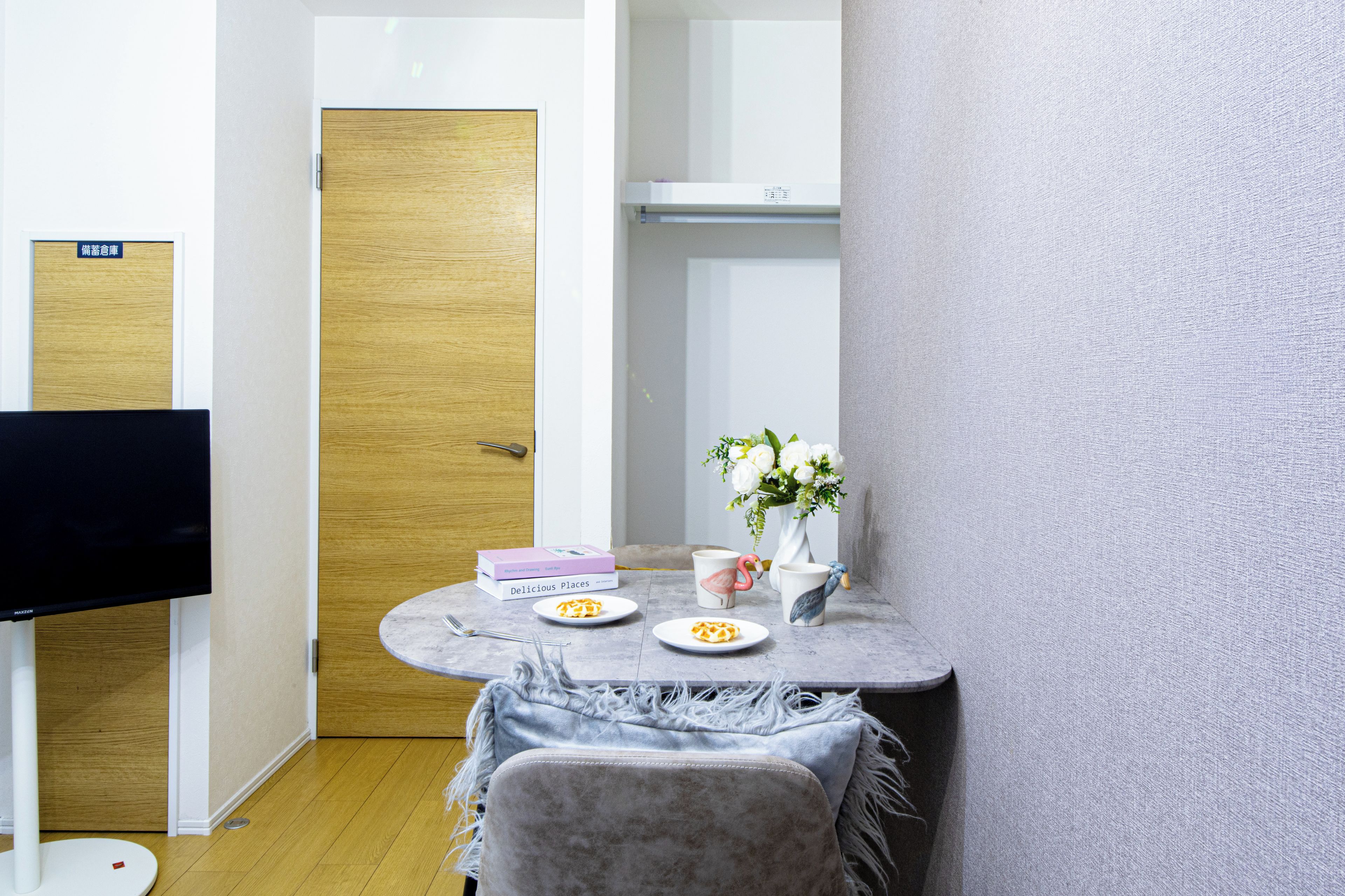 A small, neatly arranged room with a wooden door, a white wall, a round table with a vase of flowers, a couple of books, and a flat-screen TV.