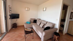 A living room with a sofa, a TV, a small table, and a washing machine in the background. - Waterfront apartment 2/3 pers. Porto-Vecchio (Porto-Vecchio)