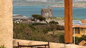 A stone structure with a wooden pergola overlooking a coastal area with a sea view, a castle-like tower, and a yellow building with solar panels.