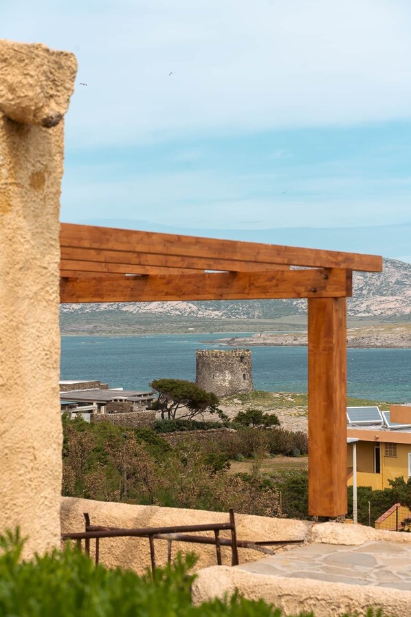 A stone structure with a wooden pergola overlooking a coastal area with a sea view, a castle-like tower, and a yellow building with solar panels.