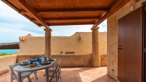 A covered outdoor dining area with a table set for two, a view of the sea, and a rustic building in the background.