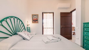 A bedroom with a green metal headboard, a bed with white linens, a folded towel, a painting on the wall, and a door leading to another room.
