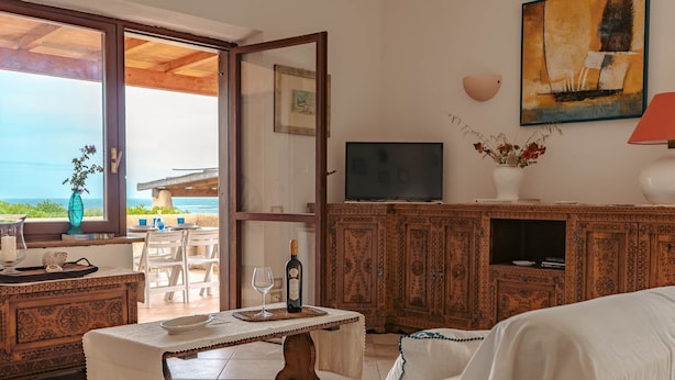 A room with a wooden cabinet, a television, a table with a bottle and glass, and a view of the sea through a glass door.