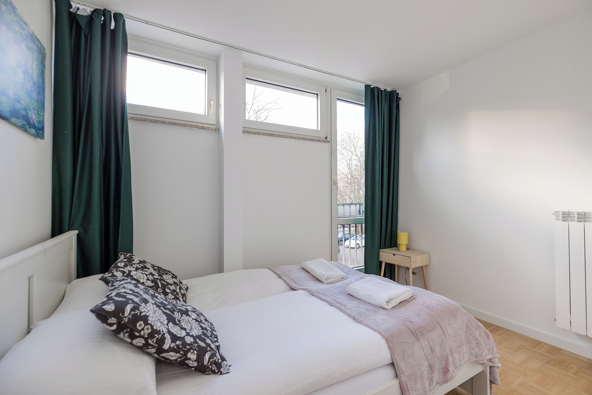 A bedroom with a bed, two windows, a radiator, and a small table with a cup.