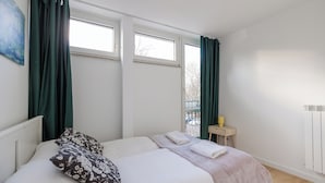 A bedroom with a bed, two windows, a radiator, and a small table with a cup. - ShortStayPoland Waliców B171 (Warsaw)