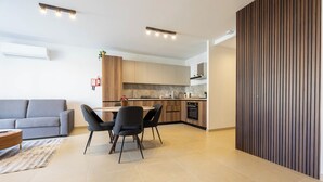 A modern kitchen with a dining area, grey sofa, and a wooden accent wall. - Cozy Living 2 BR Apartment Walk to Sea (San Pawl il-Baħar)