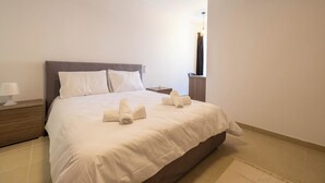 A neatly made bed with white linens and towels, a bedside table with a lamp, and a wall-mounted air conditioner. - Cozy Living 2 BR Apartment Walk to Sea (San Pawl il-Baħar)