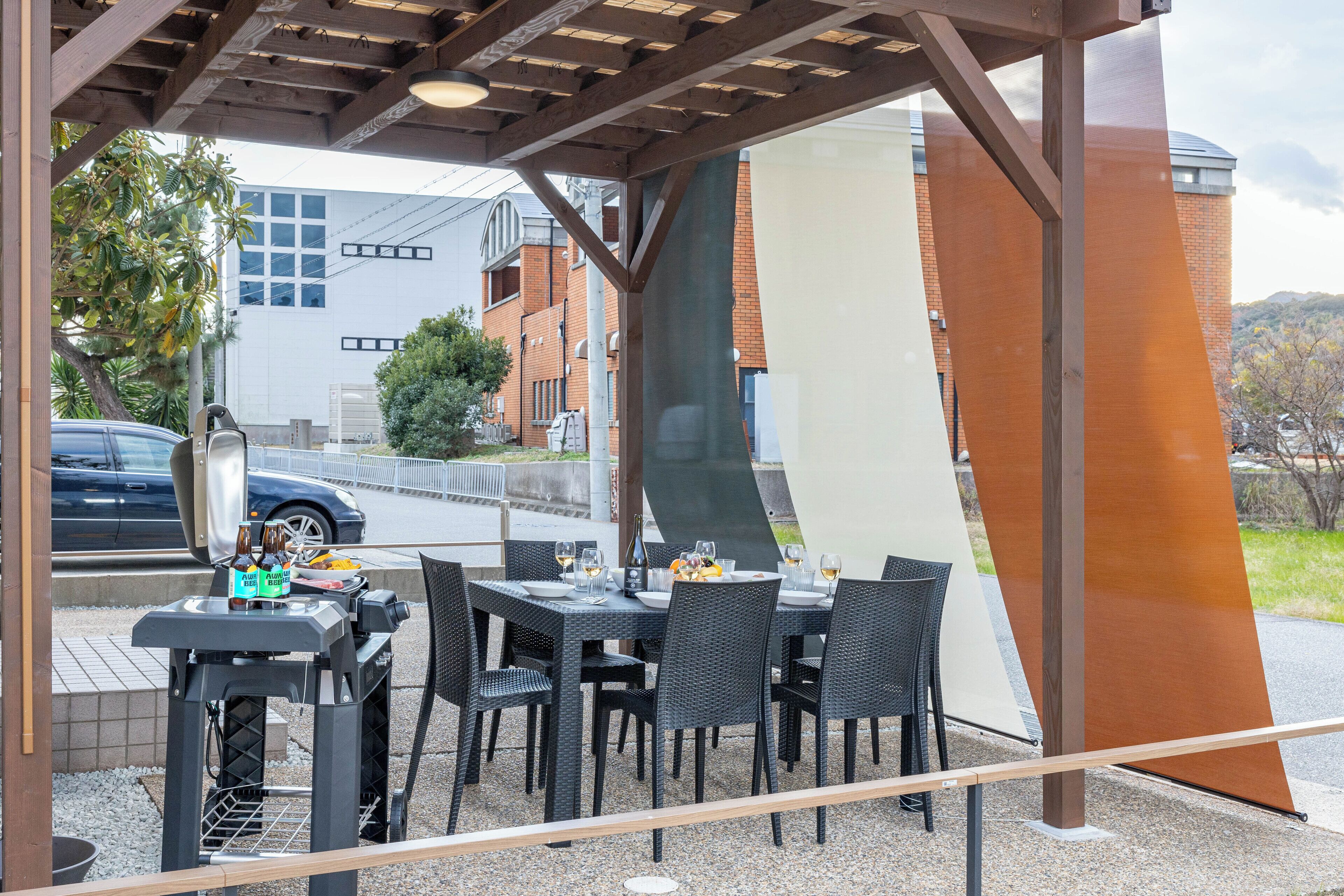 Un espacio al aire libre con mesas y sillas, una parrilla, y vista a edificios y un coche en el fondo.