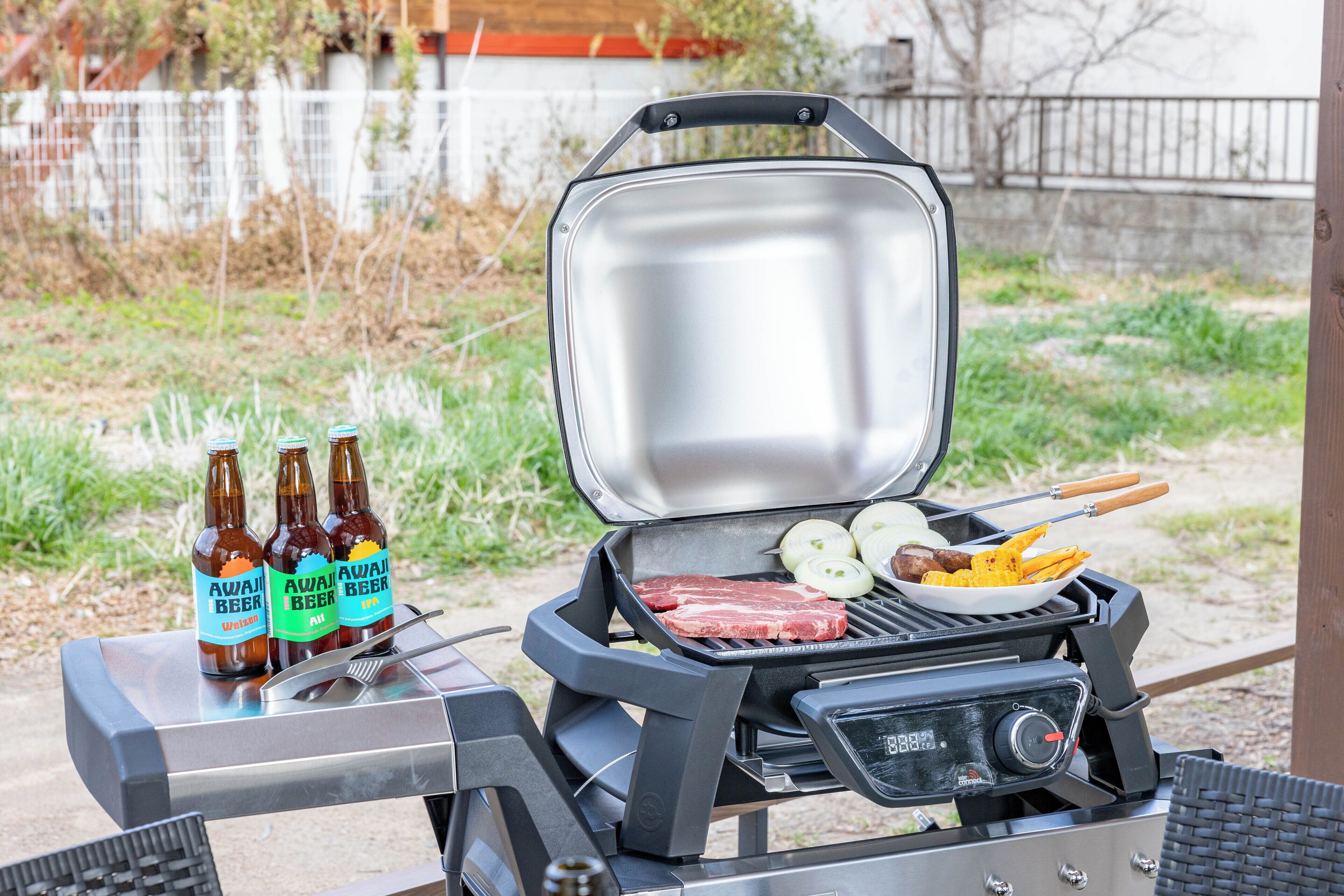 A portable grill with steaks, corn, and a side dish, and three bottles of beer on a table.