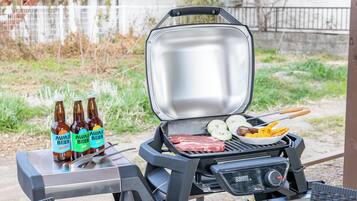 A portable grill with steaks, corn, and a side dish, and three bottles of beer on a table.