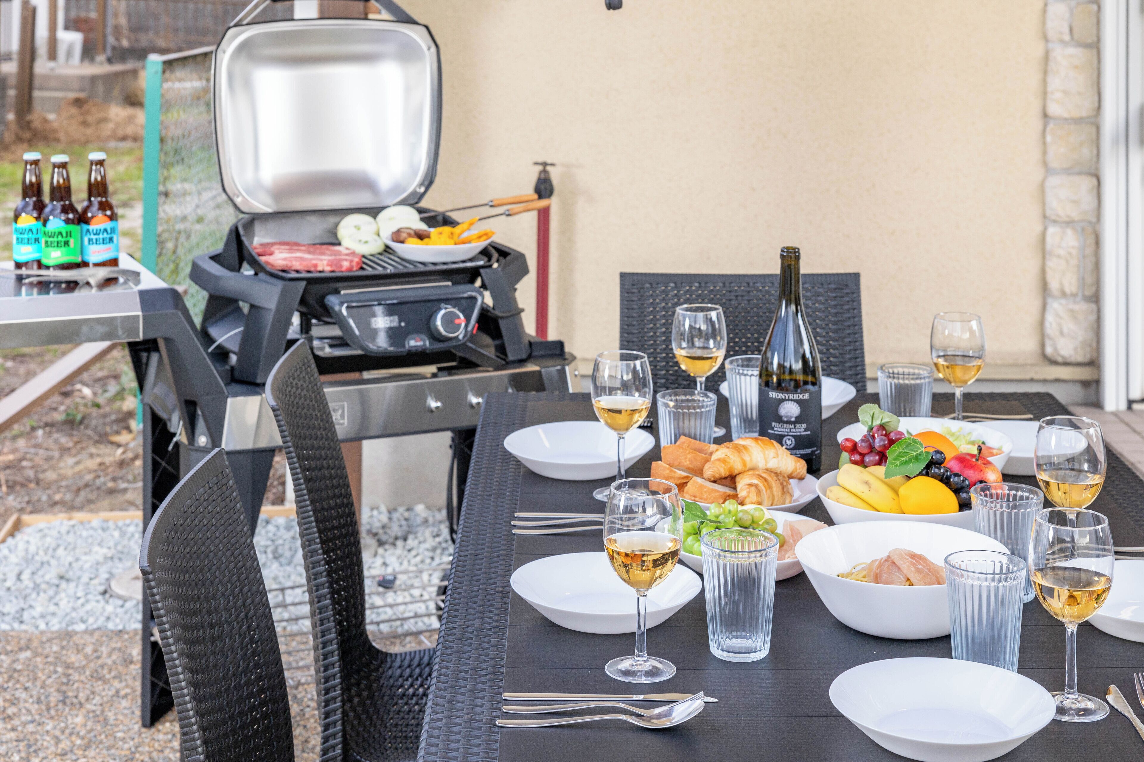 Et spisebord dækket med tallerkener, glas og flasker, en grill med mad samt en vinflaske.