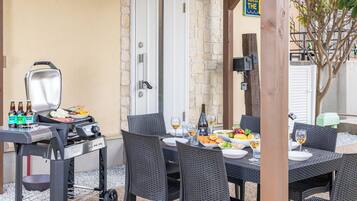 An outdoor dining area with a grill, wicker chairs, a table set with food and drinks, and a stone wall in the background.
