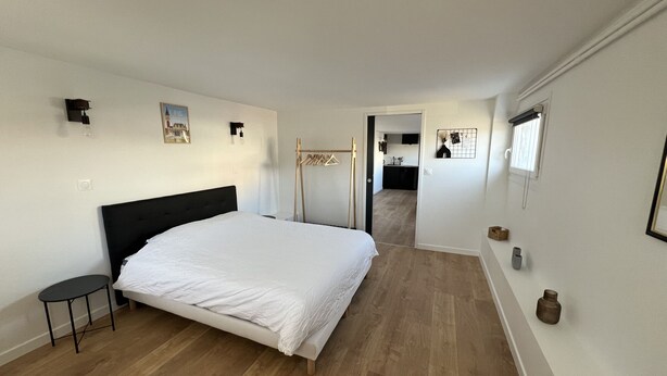 A bedroom with a large bed, a small table, a wooden floor, and a view into the kitchen. - Le Calais Wilson 4 : 2-4 Pers (Calais)