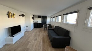 A modern living room with a flat-screen TV, a black sofa, and a wooden coffee table. There is a kitchen area with dark cabinets and a dining table. - Le Calais Wilson 4 : 2-4 Pers (Calais)