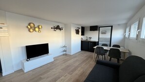 A modern living room with a flat-screen TV, a sofa, a dining area with chairs, and a kitchen with a sink and cabinets. - Le Calais Wilson 4 : 2-4 Pers (Calais)