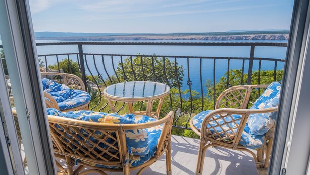 Un balcon doté de meubles en osier et d’une table en verre, offrant une vue sur une étendue d’eau et sur la terre ferme au loin.