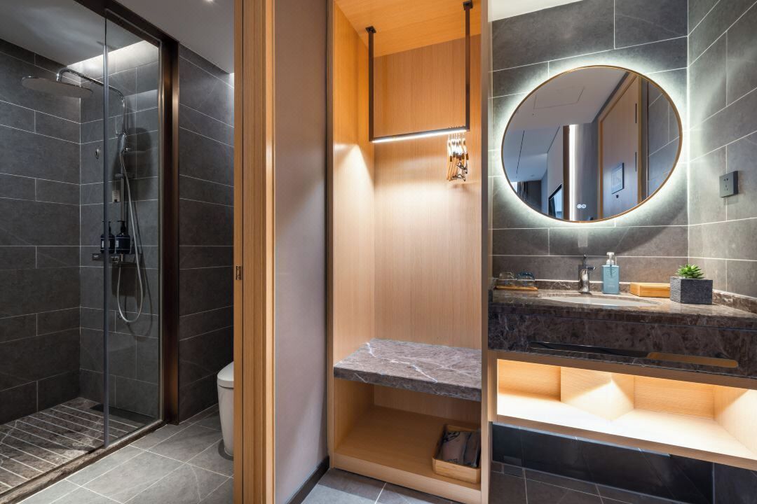 A modern bathroom with a glass-enclosed shower, a wooden vanity with a marble countertop, and a round mirror with a light.