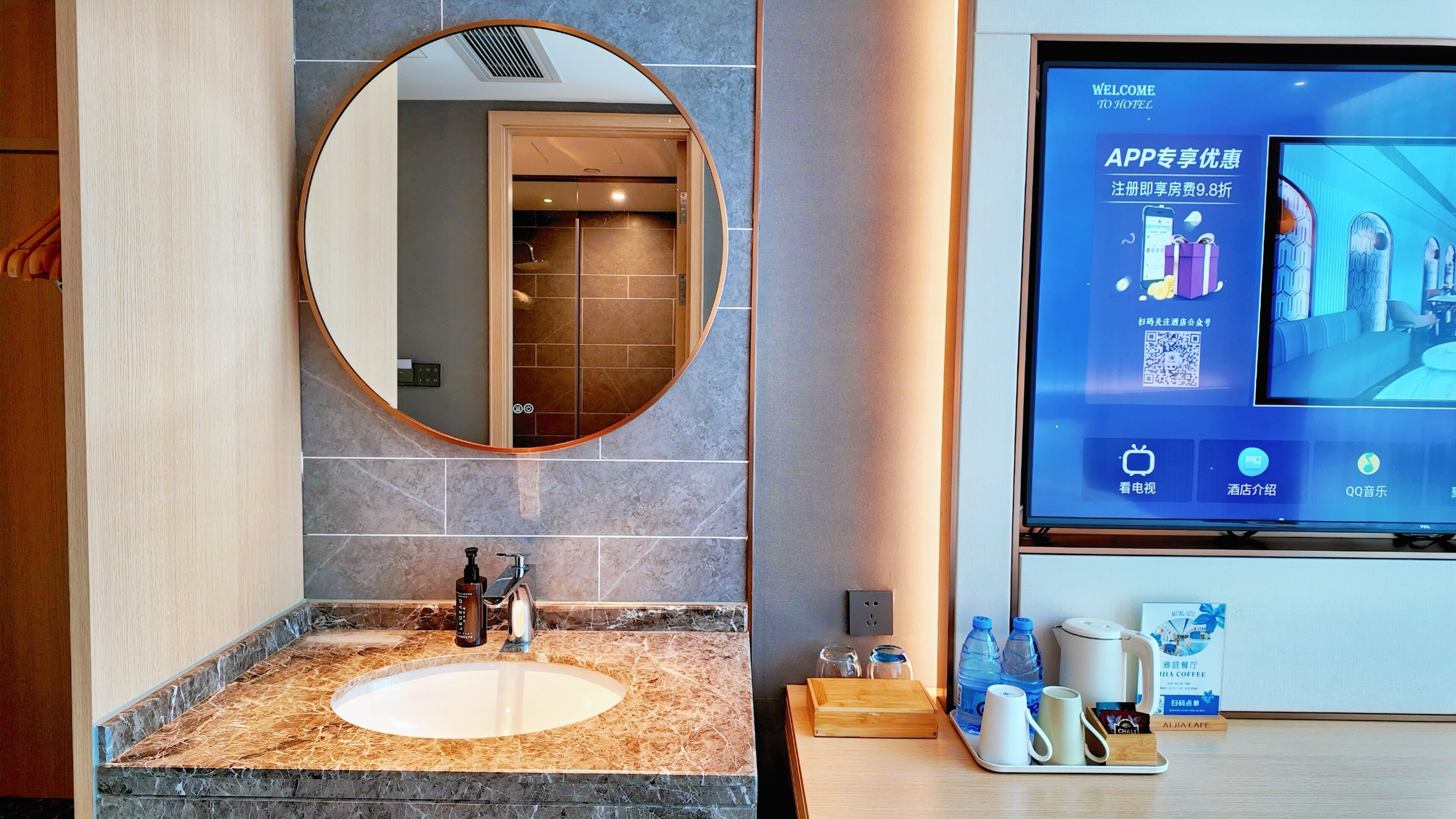 A modern bathroom with a marble sink, a round mirror, and a digital screen displaying hotel information.
