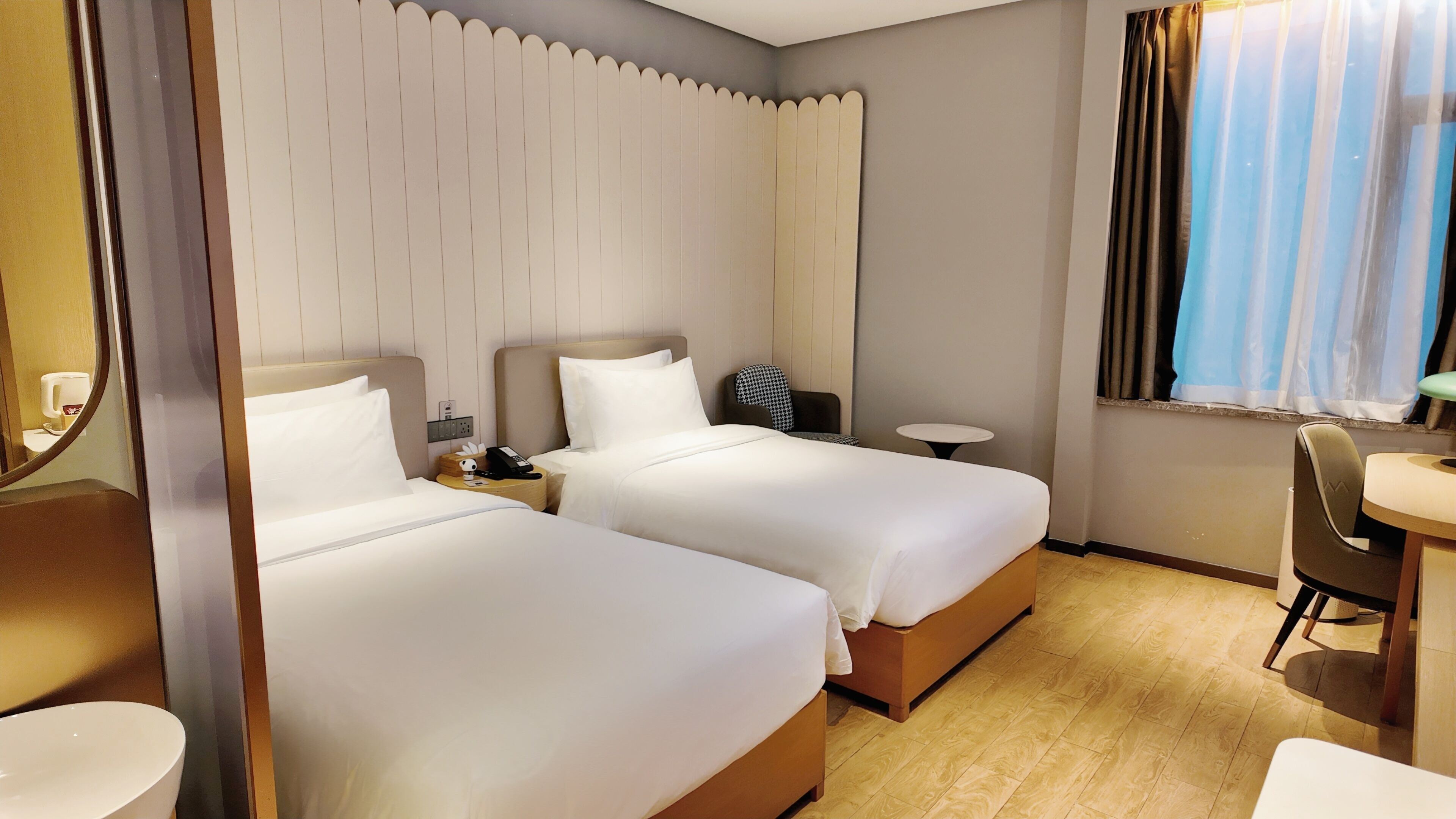 A hotel room with two beds, a small table, a chair, a mirror, and a window with curtains.