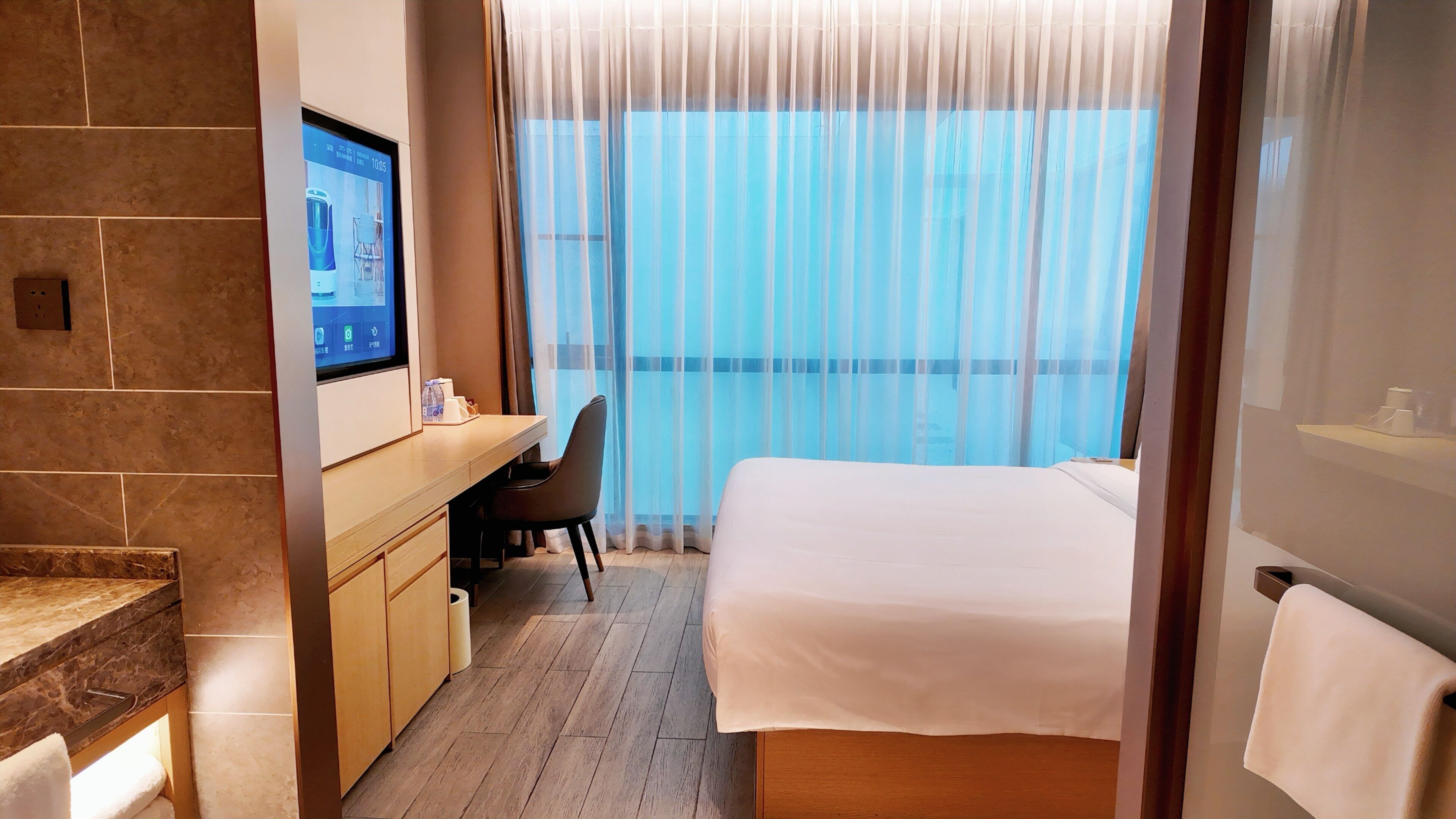Una habitación de hotel con cama, escritorio, silla, televisor y ventana con cortinas traslúcidas.