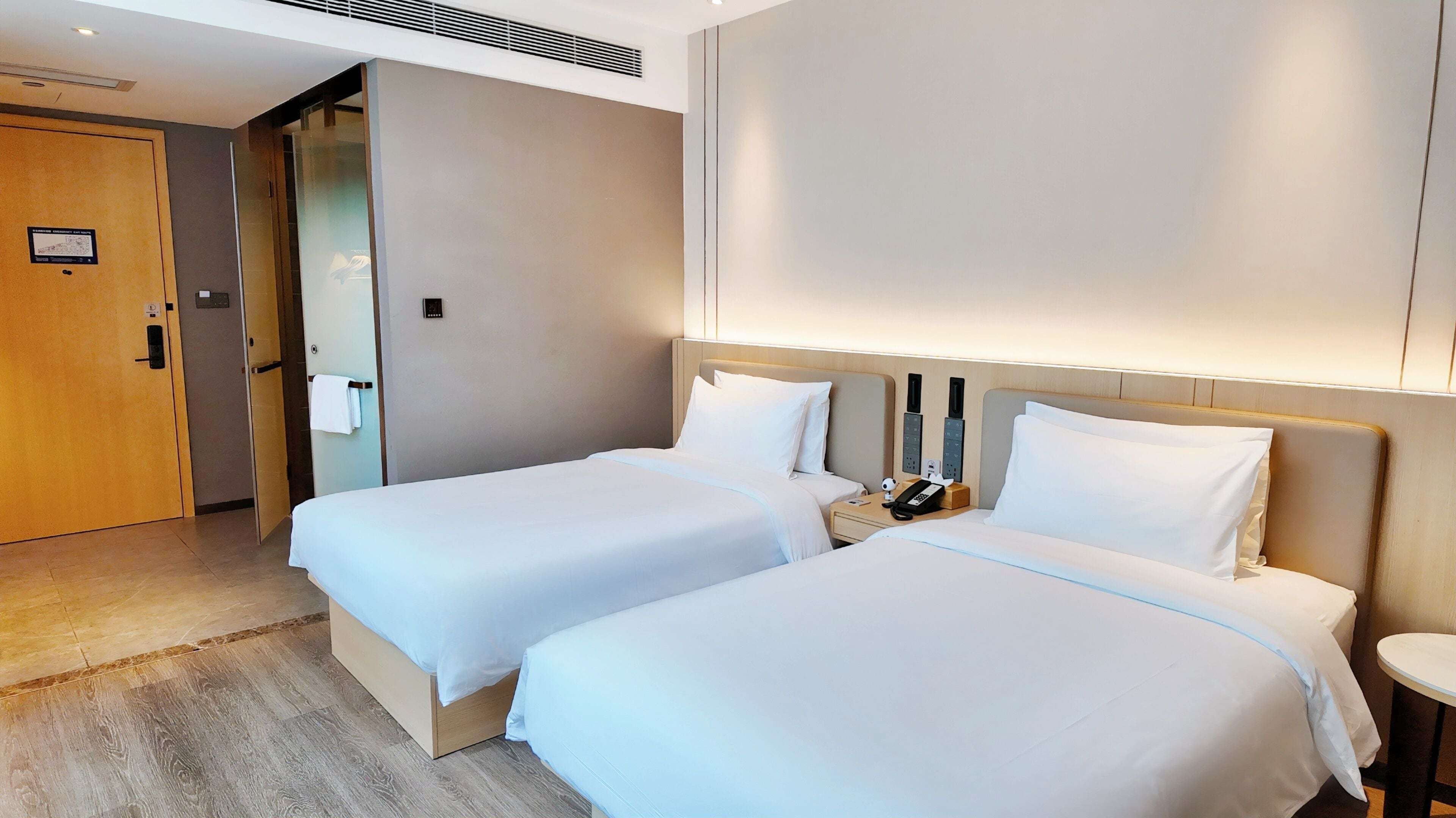 A hotel room with two beds, a wooden door, a wall-mounted control panel, and a bedside table with a phone and a lamp.