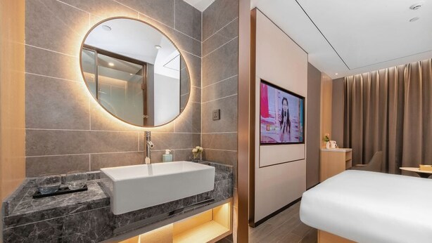 Ein modernes Hotelzimmer mit Waschtisch aus Marmor, einem großen Spiegel, einem Flachbildfernseher, einem Bett und einem kleinen Tisch.