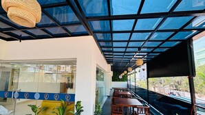 Un espacio de estar al aire libre cubierto con mesas y sillas, plantas en macetas y una pared de vidrio con una franja azul.