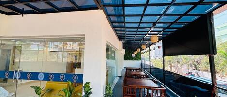 A covered outdoor seating area with tables and chairs, potted plants, and a glass wall with a blue stripe.