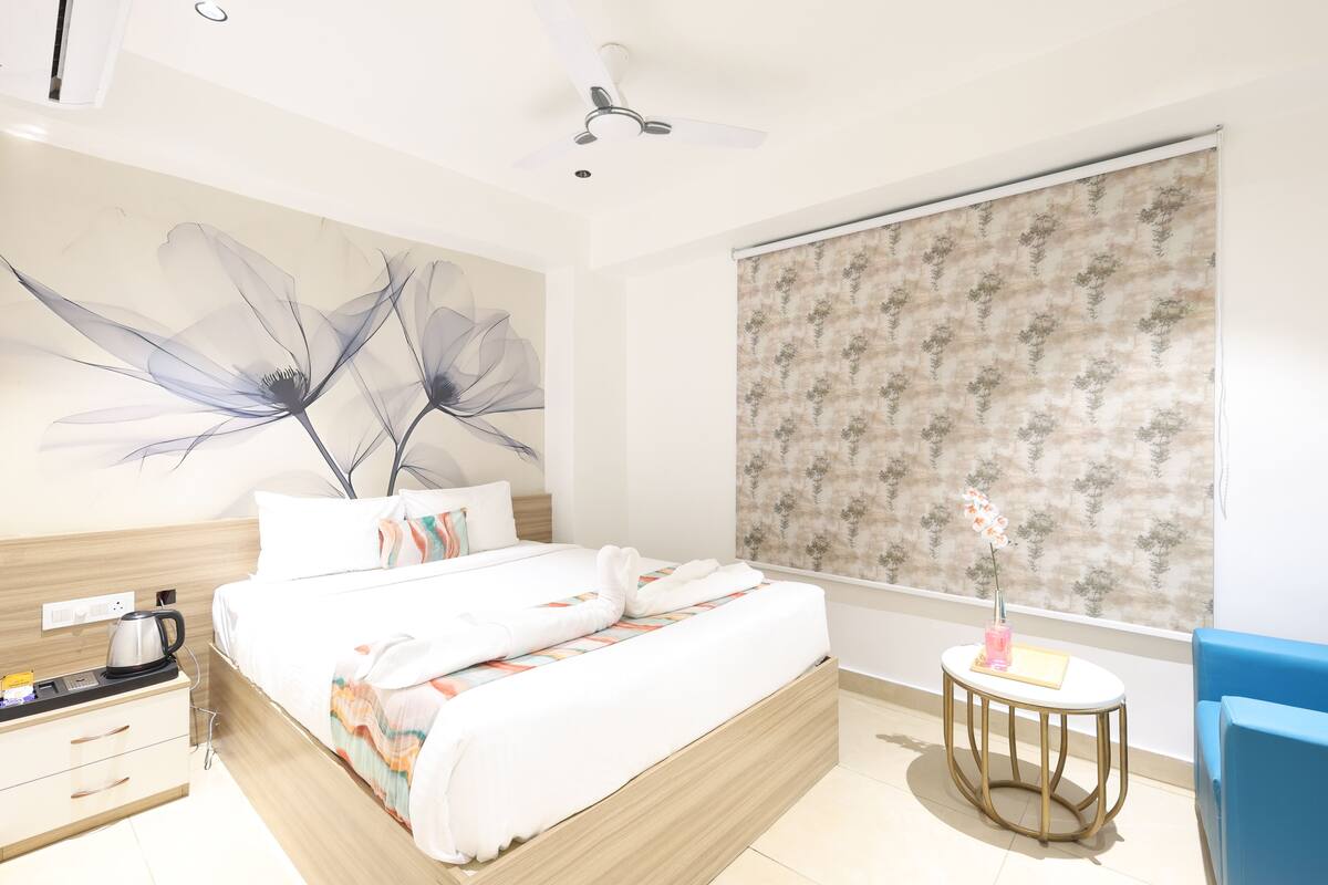 Une chambre d’hôtel avec un lit, une table ronde ornée d’un vase, une chaise bleue, un ventilateur de plafond et une grande fenêtre avec un rideau à motifs floraux.