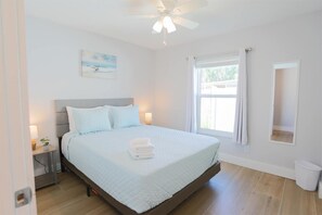 Quarto com cama, ventoinha no teto, janela com cortinas, mesa de cabeceira com abajur e pintura pendurada na parede.