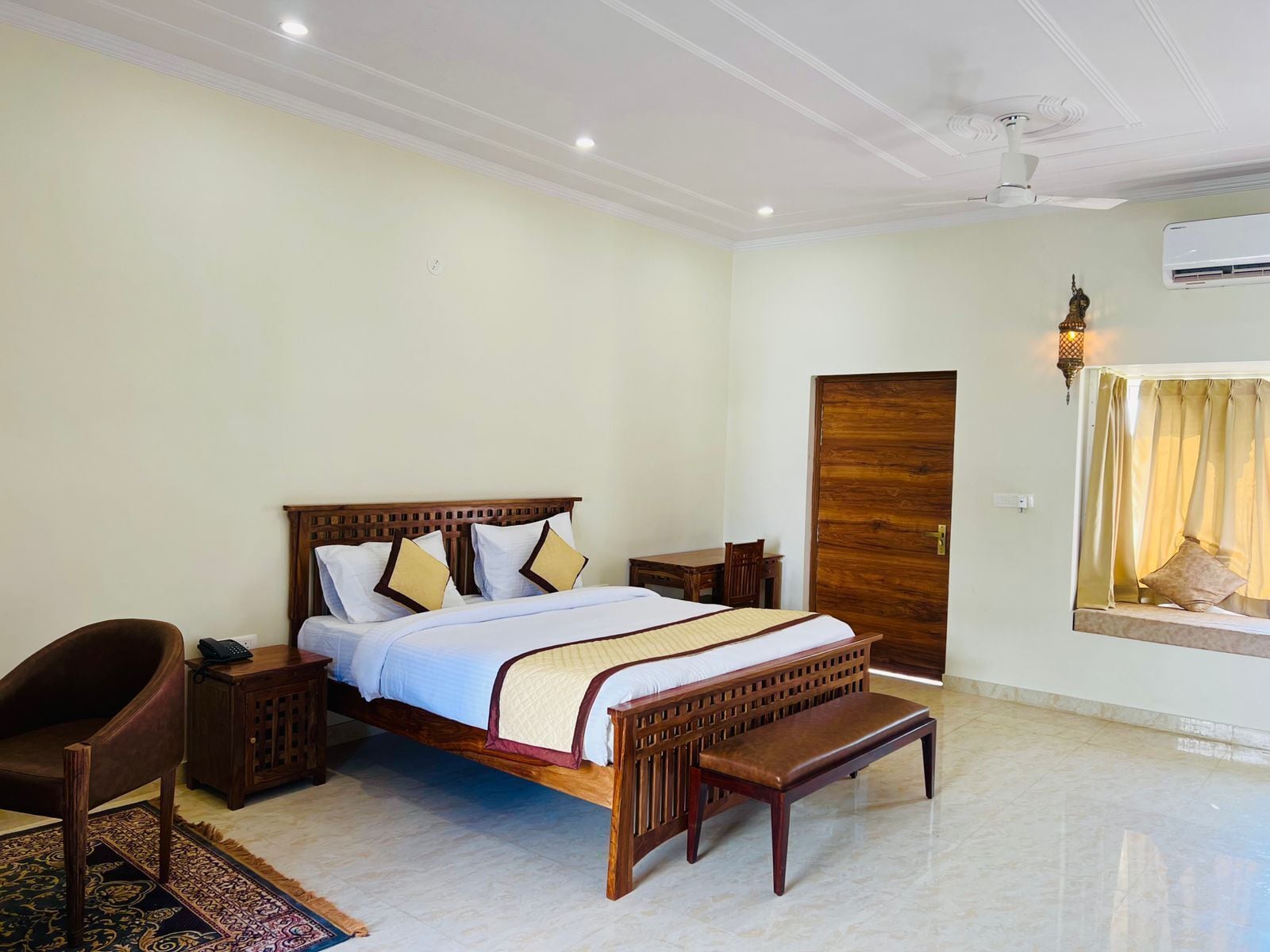 Une chambre à coucher avec un lit en bois, des tables de chevet, un banc, une chaise, un tapis à motifs et une fenêtre avec des rideaux.