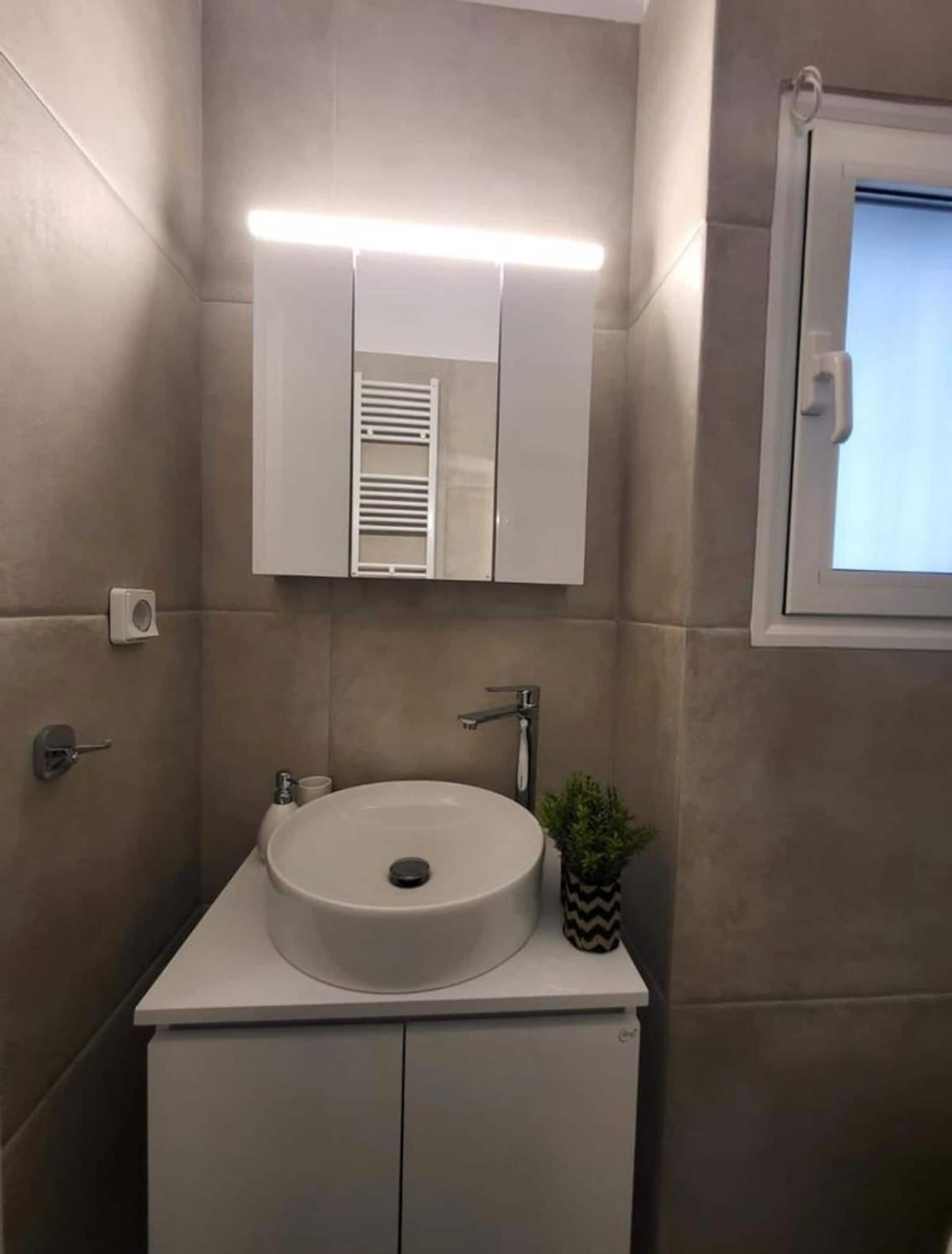A modern bathroom with a round sink, a mirror above it, and a window to the right.