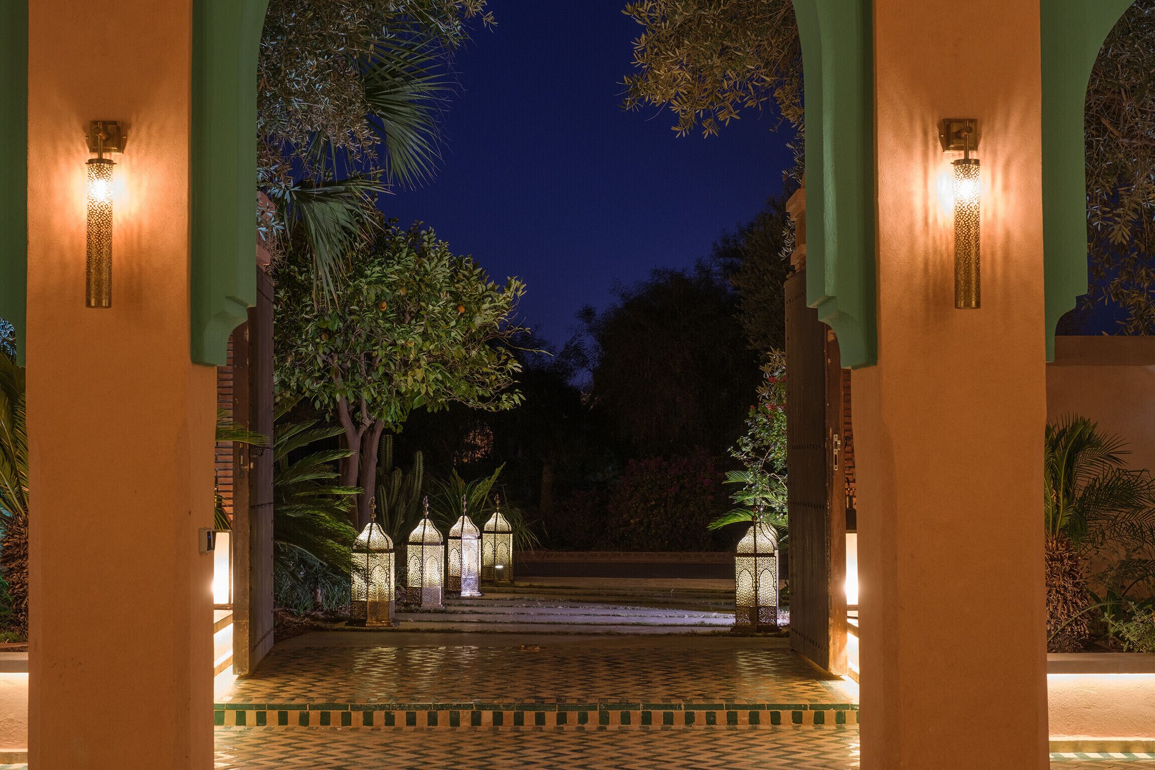 Vista nocturna de un patio con piso a cuadros, palmeras y faroles iluminados.