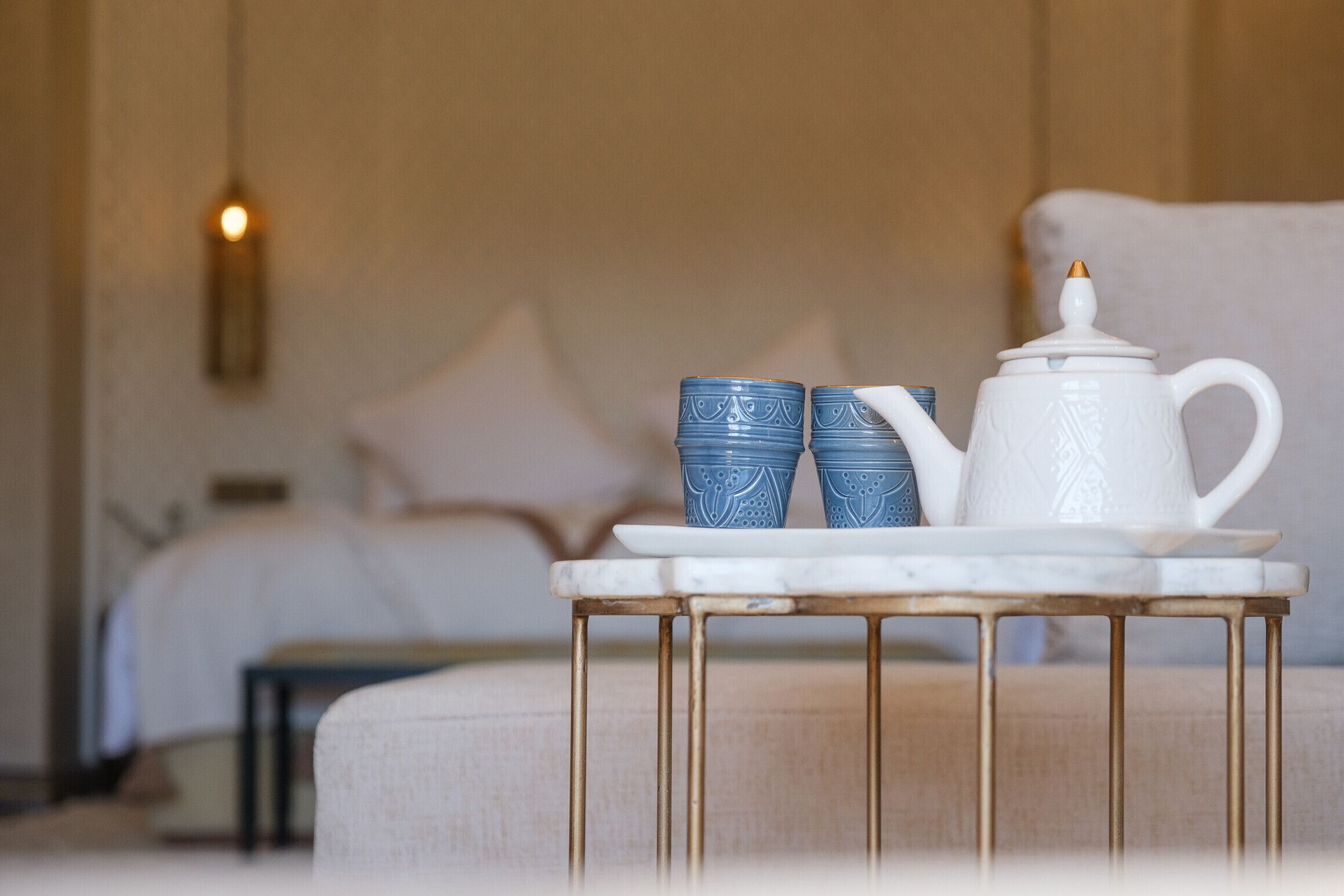 A neatly arranged table with a white teapot and two blue cups on a marble surface, set against a softly lit room with a bed and a wall-mounted light.
