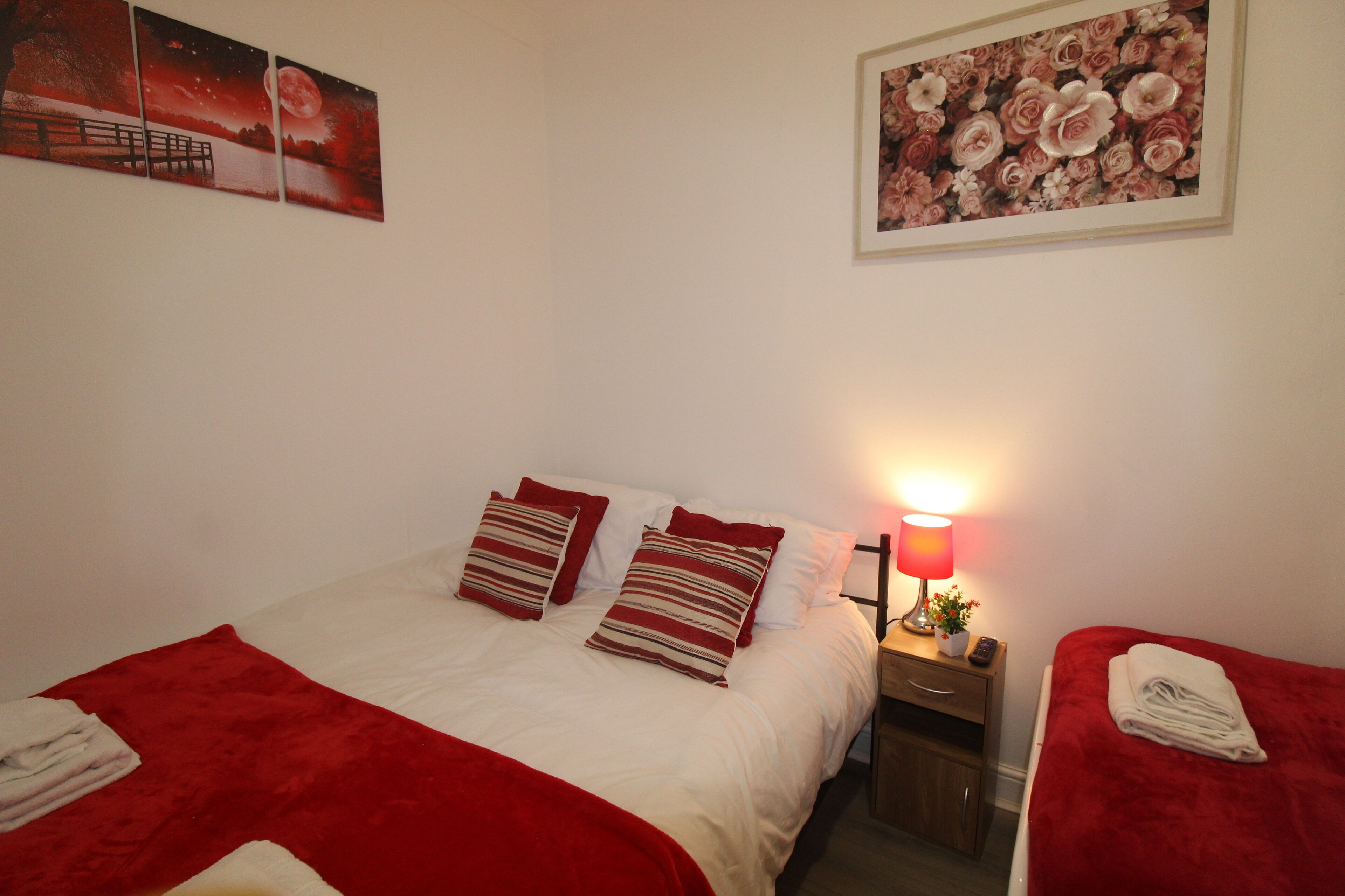A hotel room with a bed, two pillows, a nightstand, a lamp, a red blanket, and a wall with two framed pictures.