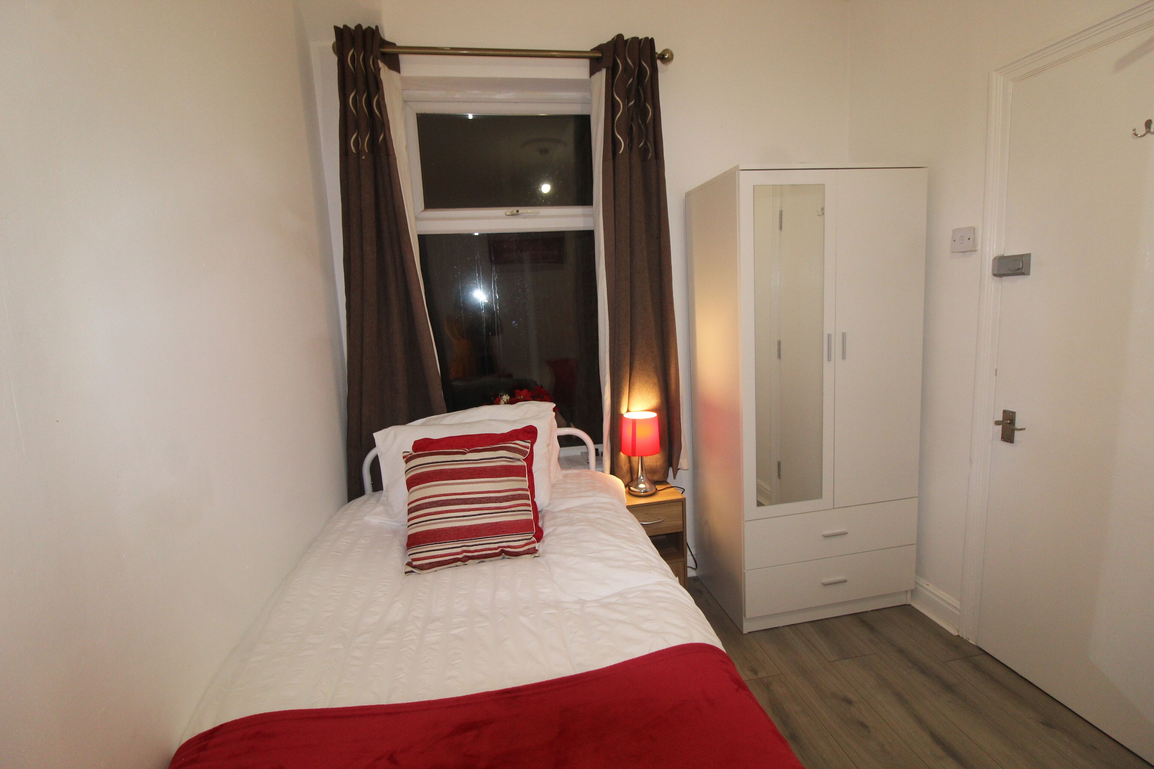 A bedroom with a bed, a nightstand with a lamp, a wardrobe, and a window with curtains.