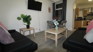 Un salon avec deux canapés noirs, une table basse en bois, une petite table d’appoint avec une plante, un téléviseur fixé au mur et une vue sur la cuisine par une porte ouverte.