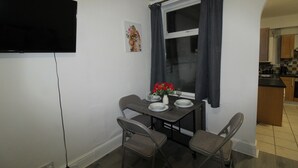 Un petit coin repas avec une table dressée pour deux personnes, une fenêtre avec des rideaux, un téléviseur à écran plat fixé au mur et une cuisine visible à travers une arche.