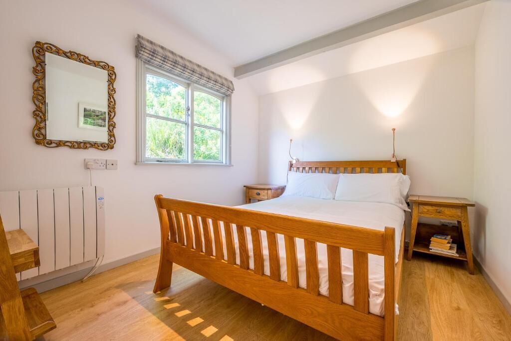 A bedroom with a wooden bed, a bedside table, a nightstand, a mirror, and a window with a view of greenery.