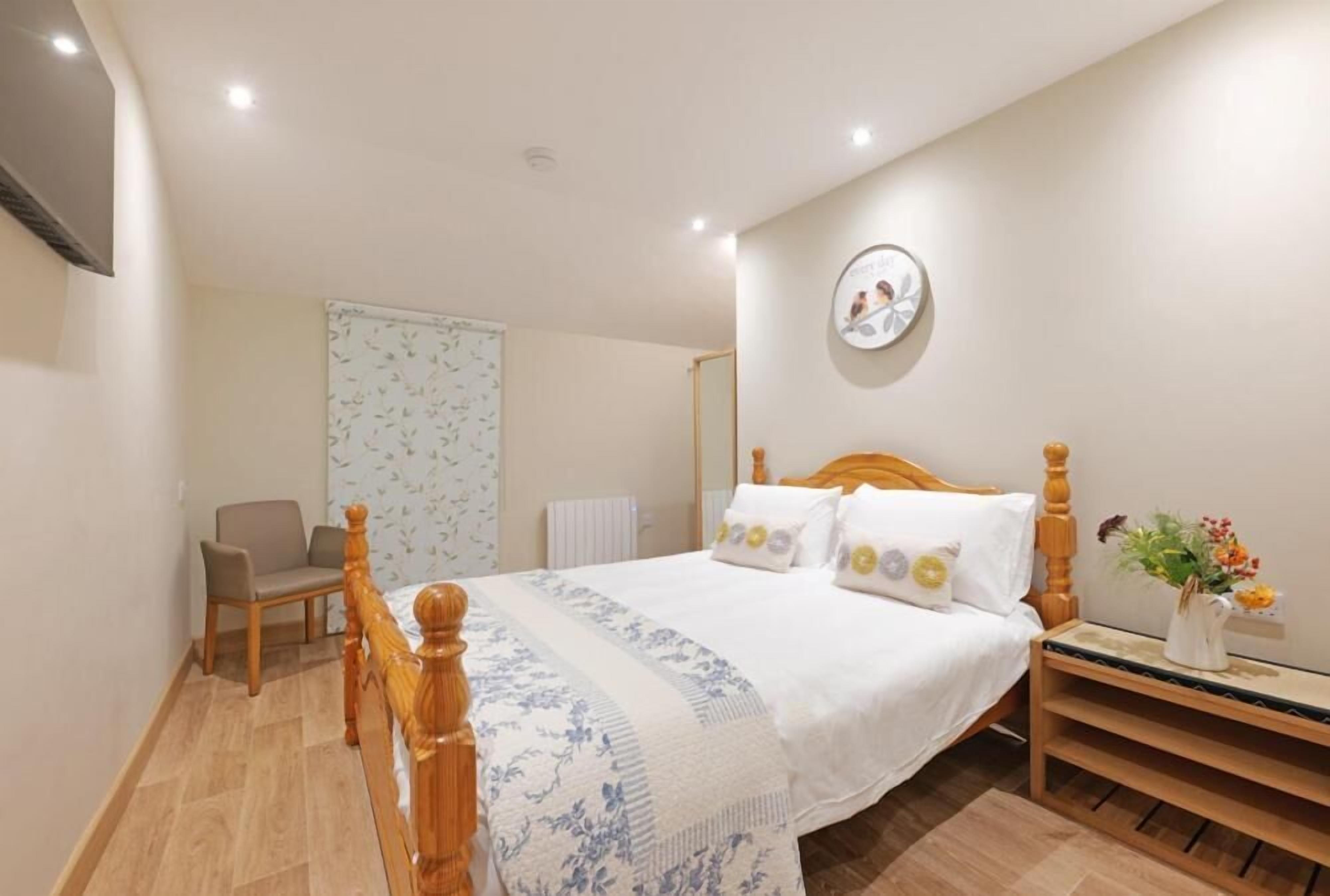 A bedroom with a wooden bed, a chair, a bedside table with a vase of flowers, and a wall clock.