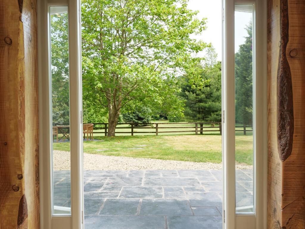 Uitzicht door een houten deur op een stenen terras, met een houten hek en bomen op de achtergrond.