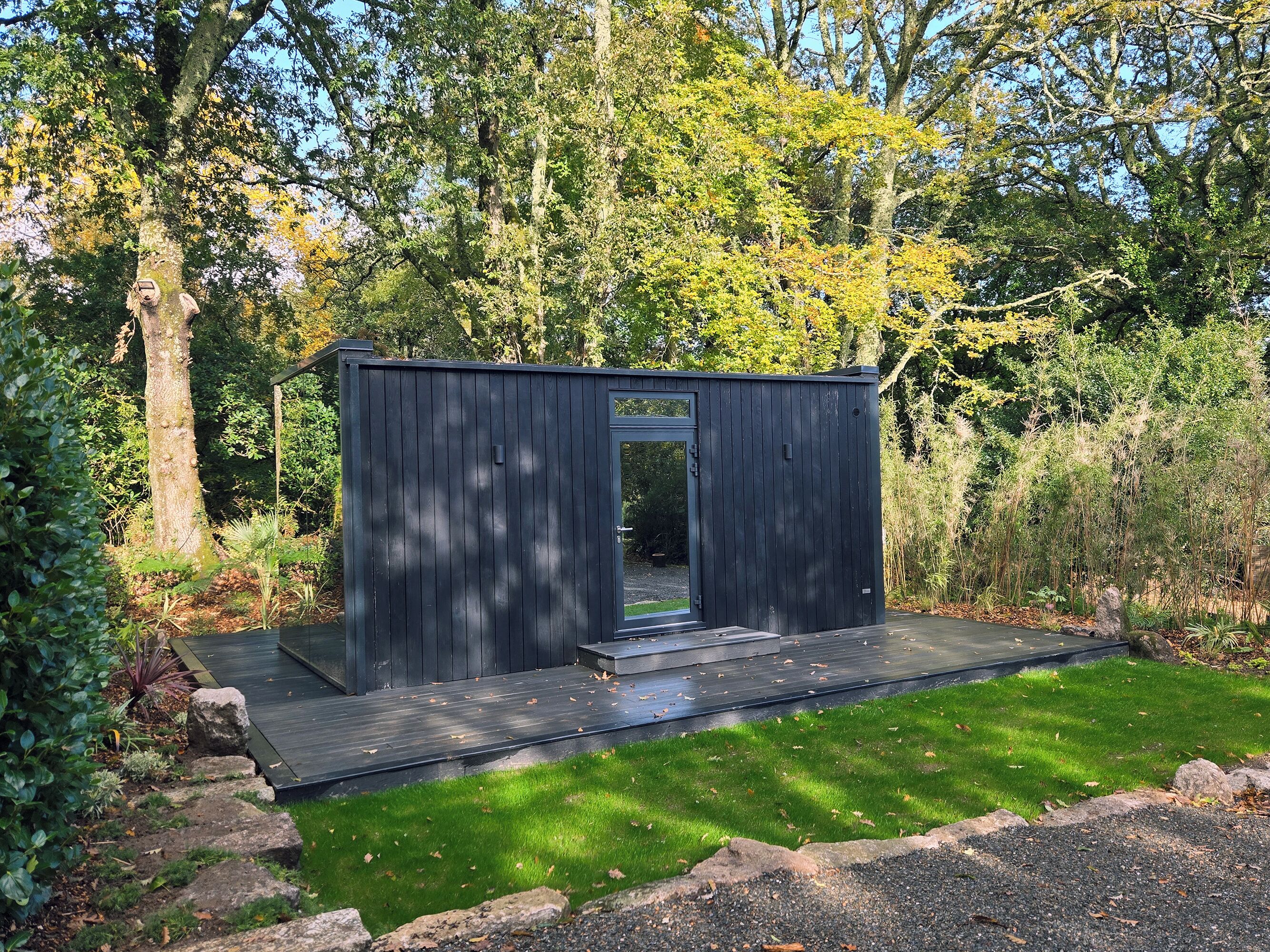 A small, dark wooden structure with a glass door, surrounded by a well-maintained lawn and dense foliage.