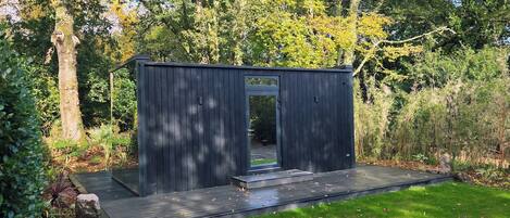 A small, dark wooden structure with a glass door, surrounded by a well-maintained lawn and dense foliage.
