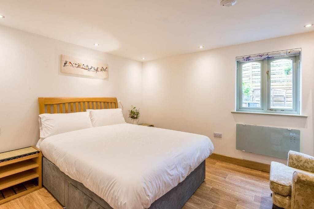 A bedroom with a large bed, a wooden headboard, a nightstand with a lamp, a window with blinds, and a radiator.