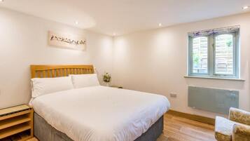 A bedroom with a large bed, a wooden headboard, a nightstand with a lamp, a window with blinds, and a radiator.