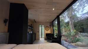 Un dormitorio moderno con una cama grande, un escritorio pequeño y una silla. Hay una ventana con vistas a árboles y un reloj en la pared.