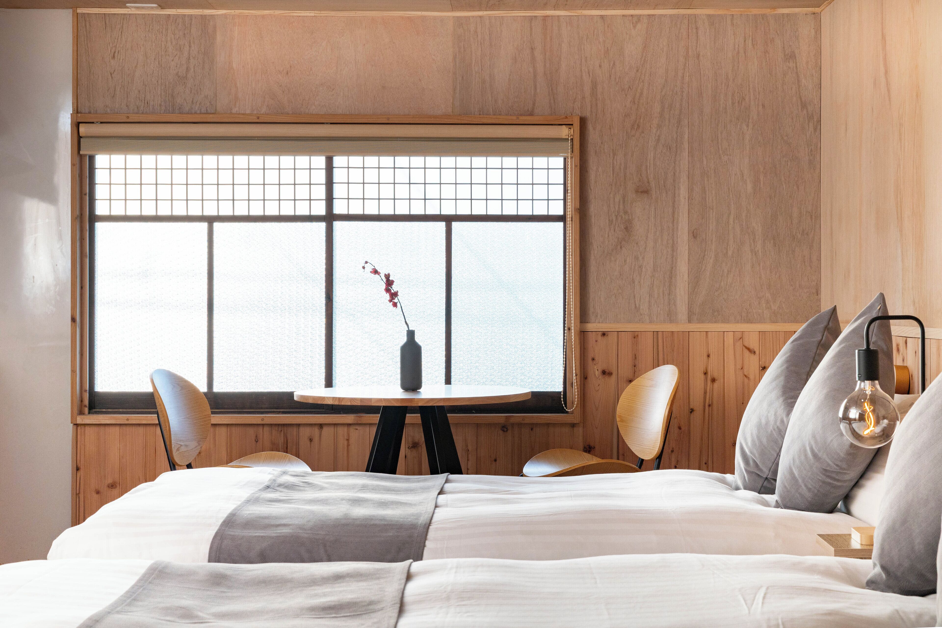 A bedroom with a large window, a round table, two chairs, a bed, and a bedside lamp.
