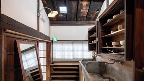 A traditional Japanese-style room with tatami flooring, a sliding shoji screen, a wooden shelf with dishes, and a stone sink. - Mameyama15 (Nara)